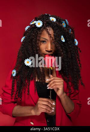 Lebhaftes Porträt einer jungen Afroamerikanerin in einem leuchtend roten Jackett, die eine rote Rose hält. Ideal für Beauty-, Mode- und Lifestyle-Branding mit romantischem oder elegantem Touch. Stockfoto