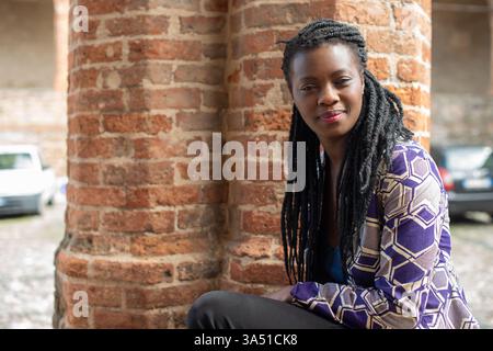 Fröhliche junge Schwarze Frau sitzt im Freien in einem modischen Look mit geflochtenen Dreadlocks. Dieses Porträt mit selektivem Fokus passt zu Moden, Lifestyle und Empowerment. Stockfoto