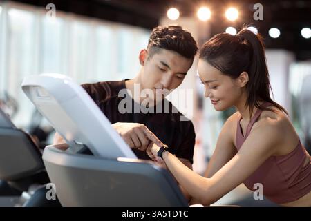 Ein chinesischer Fitnesstrainer führt eine Kundin während eines Fitnesstrainings und passt ihre Smartwatch an. In diesem Moment werden tragbare Technologien und persönliches Coaching in einer modernen Fitness-Umgebung vorgestellt. Ideal für Wellness-, Fitness- und Sportswear- oder Fitnessgeräte-Aktionen. Stockfoto