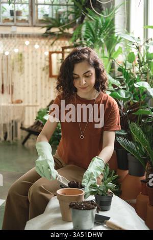 Eine junge Frau in lässiger Kleidung pflanzt eine grüne Pflanze in einen Topf im Garten. Sie sitzt draußen, lächelt, während sie sich um Zimmerpflanzen kümmert und ein entspannendes Hobby genießt. Ideal für Inhalte über Gartenarbeit, das Leben zu Hause und die Natur. Stockfoto