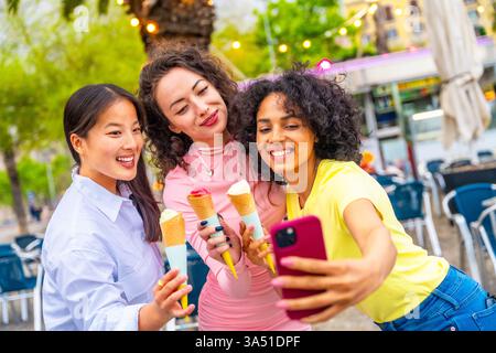 Drei verschiedene Freundinnen machen ein Selfie, während sie im Urlaub Eis genießen. Die unbeschwerte, sonnige Atmosphäre unterstützt Reisen, Freundschaften und Lifestyle-Kampagnen im Sommer. Ideal für soziale Medien, Tourismus und Jugendmarketing. Stockfoto