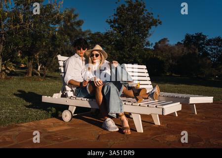 Ein junges hispanisches Paar sitzt an einem sonnigen Tag dicht beieinander auf einem Liegestuhl. Sie wirken entspannt und verbunden und genießen Outdoor-Freizeit in einer warmen Umgebung. Verwenden Sie dieses Bild für Romantik, Beziehungen und unbeschwerte Sommermomente. Stockfoto