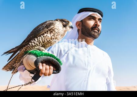 Mann aus dem Nahen Osten in einem traditionellen Emirat-Kandura, der einen Falken hält, während er in Wüstendünen steht. Authentisches Porträt, geeignet für Kultur des Nahen Ostens, Falknerei und geschäftliche oder kulturelle Themen der VAE. Ideal für Reisen, Tourismus oder Geschäftsreisen in der Golfregion. Stockfoto
