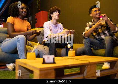 Lächelnde hispanische weibliche und männliche Freunde sitzen auf der Couch und spielen Videospiele in der Computerhalle Stockfoto