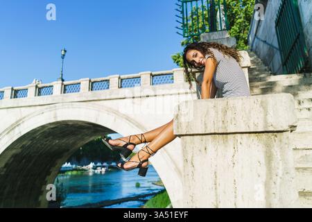 Die lächelnde hispanische Frau sitzt auf Beton nahe einer Brücke in einer städtischen Tagesumgebung. Die durchdachte Pose ruft Reisen, Erkundungen und ungezwungene Stadtatmosphäre hervor. Ideal für Reisen, Tourismus und Lifestyle-Kampagnen mit urbanen Landschaften. Stockfoto
