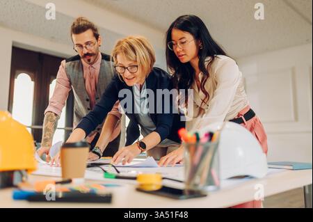 Verschiedene Architektengruppen arbeiten gemeinsam an Bauplänen auf einem Tisch im Büro. Sie stehen zusammen, analysieren Pläne und tauschen Ideen in einem kreativen Arbeitsbereich aus. Perfekt für Geschichten über Design, Architektur, Teamarbeit und Zusammenarbeit im Büro. Stockfoto