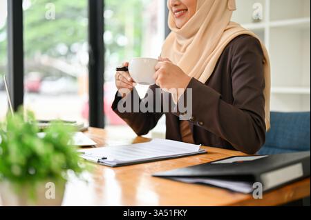 Nahaufnahme einer asiatischen muslimischen Geschäftsfrau, die einen Hijab trägt und an ihrem Schreibtisch in einem modernen Büro Kaffee trinkt. Selbstbewusstes, professionelles Porträt, das sich für Unternehmenskommunikation und das Branding bei der Vielfalt am Arbeitsplatz eignet. Ideal für Geschäfts- und Führungsmaterialien. Stockfoto