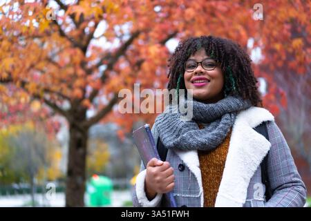 Ein lächelnder hispanischer Student mit lockigen Haaren steht im Herbst draußen und hält eine Mappe in der Nähe von Campus-Bäumen. Ein ideales Bild für Schulanfänger, College- und Outdoor-Studienszenen, das Optimismus und Lernenergie vermittelt. Stockfoto