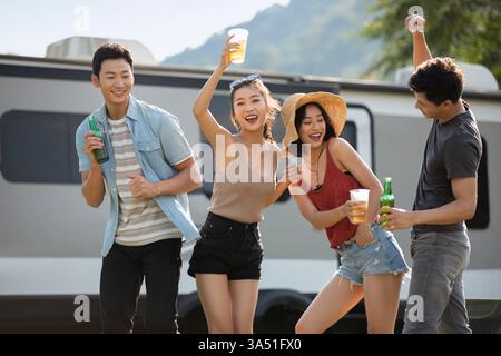 Lächelnde chinesische Freunde tanzen in einem sonnigen Park in der Nähe eines Wohnwagens. Sie heben Gläser und Flaschen auf und feiern mit einer entspannten Picknickatmosphäre. Ideal für Reisen, Freundschaften und Outdoor-Lifestyle-Aktionen. Stockfoto