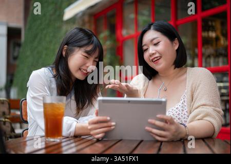 Zwei fröhliche asiatische Freunde sitzen an einem Coffee Shop-Tisch im Freien, plaudern und teilen ihre Meinungen. Sie lächeln in einem hellen Café, perfekt für Momente der Freundschaft und zwangloser Unterhaltung. Stockfoto
