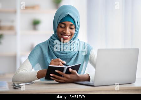Schwarze muslimische Frau mit Hijab arbeitet an einem Schreibtisch mit Laptop und Notizblock in einem Heimbüro. Sie wirkt fokussiert und stellt freiberufliche und Remote-Arbeiten in einem professionellen Umfeld dar. Stockfoto