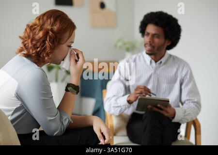Fesselndes Porträt einer jungen Frau während einer Therapiesitzung, sichtbar emotional, während sie spricht. Dieses Bild verankert Kampagnen zur psychischen Gesundheit oder Beratungskommunikation, mit einem professionellen Büro-Setting und Hinweisen von Emotionen, Diskussionen und Unterstützung. Stockfoto