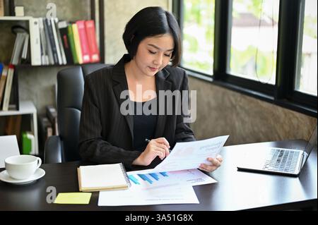 Professionelle Millennial-asiatische Führungskraft an ihrem Schreibtisch, die Marketingberichte für eine neue Kampagne durchliest. Fokussierte, selbstbewusste Bilder, die sich für Business-, Führungs- und Marketing-Branding eignen. Büroeinrichtung mit professioneller Kleidung und strategischer Planung. Stockfoto