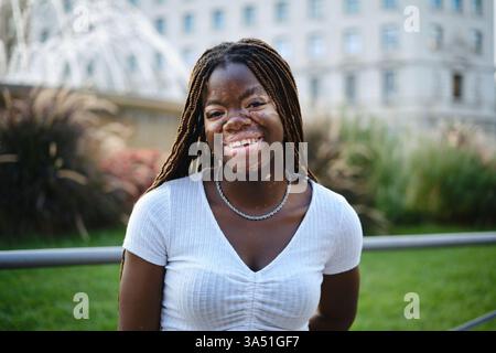 Lächelnde schwarze Frau mit Vitiligo, die bei Sonnenuntergang auf einem Stadtparkrasen steht. Ein strahlendes Porträt im Freien, das Vielfalt und Selbstvertrauen zelebriert. Stockfoto