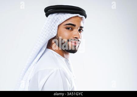 Lächelnder Mann aus dem Nahen Osten, der Kandura trägt und über die Schulter blickt, während er vor weißem Hintergrund im Studio steht Stockfoto