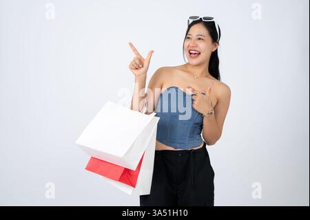 Asiatische Frau in trendiger Kleidung mit Einkaufstaschen auf einem isolierten weißen Hintergrund zeigt auf den Kopierraum. Dieses Bild eignet sich ideal für den Einzelhandel und Werbelayouts und bietet Platz für Text. Nützlich für E-Commerce-, Marketing- und Shop-Marketing-Grafiken. Stockfoto