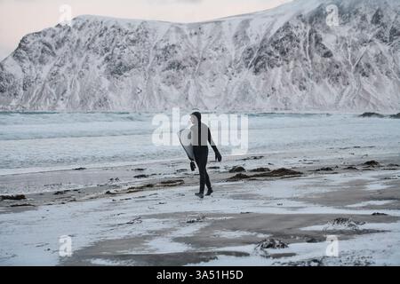 Authentische lokale arktische Surfer spazieren am Strand nach einem Winter Surfen an der Nordseeküste Norwegens. Das blaue arktische Wasser und die zerklüftete Küste werden von einer kalten, winterlichen Atmosphäre mit einem Neoprenanzug und Surfbrett umrahmt. Eine gute Wahl für Abenteuer, Extremsportarten und nordische Reisekollektionen. Stockfoto
