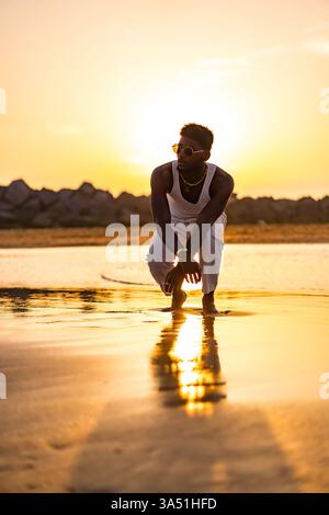 Selbstbewusster Schwarzer Mann, der sich bei Sonnenuntergang am Strand hockt, in einem weißen Tanktop und einer leichten Hose mit Sonnenbrille gekleidet. Diese Outdoor-Lifestyle-Aufnahme vermittelt eine stilvolle, entspannte Atmosphäre, ideal für Reisen, Mode und Sommerkampagnen. Perfekt für Strandbekleidung, Freizeit und unbeschwerte Stimmung in warmen Farben. Stockfoto