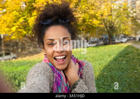 Fröhliche Afroamerikanerin macht tagsüber ein Selfie in einem sonnigen Park. Dieses helle Porträt unterstreicht ihr natürliches Lächeln, ihren selbstbewussten Ausdruck und ihren sorglosen Stil in einer Parklandschaft. Ideal für Lifestyle, Outdoor-Aktivitäten oder Selbstpflegethemen mit einer freundlichen, zugänglichen Atmosphäre. Stockfoto