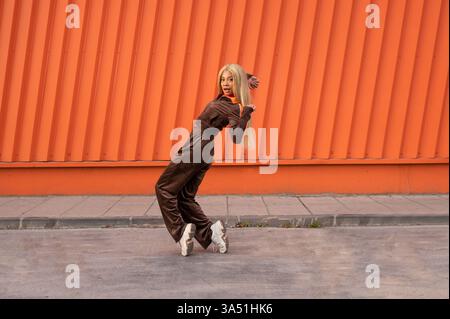 Glückliche junge Frau mit blonden Haaren in sportlicher Kleidung, die auf einer Stadtstraße tanzt. Die dynamische Pose und die helle Energie machen es zu einer idealen Wahl für Fitness-, Tanz- oder Street-Fashion-Kampagnen. Ideal für Lifestyle- und urbane Aktivitäten. Stockfoto
