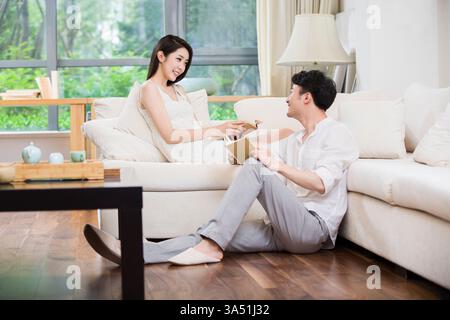 Fröhliches, junges chinesisches Paar liest ein Buch in einem komfortablen Wohnzimmer. Diese ehrliche Heimszene beleuchtet Zweisamkeit, Entspannung und häusliches Leben, perfekt für Geschichten über Paare, asiatische Haushalte und gemütliche Innenarchitektur. Stockfoto