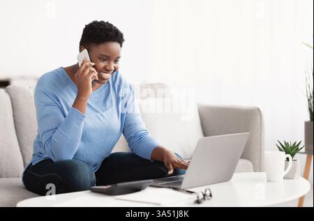 Lächelnde Afroamerikanerin arbeitet von zu Hause aus auf dem Sofa. Sie chatten auf ihrem Smartphone, während sie einen Laptop verwendet, und veranschaulicht dabei Remote-Arbeiten und ein modernes Home Office. Dieses Bild eignet sich perfekt für Lifestyle- und Business-Visuals, die Telearbeit und digitale Konnektivität betonen. Stockfoto
