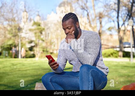 Der schwarze Mann sitzt auf einer Bank im Freien mit einer nachdenklichen Pose, die Hand liegt auf dem Kinn. Dieses urbane Lifestyle-Bild passt zu legeren Porträts und modernen Umgebungen. Stockfoto