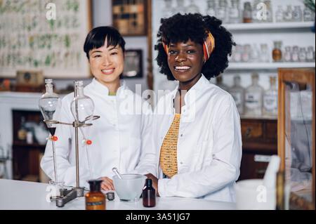 Porträt eines multirassischen Apothekers in einem Labormantel, der in einer alten Drogerie steht und neben Laborgefäßen die Kamera anlächelt. Diese authentische Pharmazie-Szene ist für das Branding in den Bereichen Gesundheitswesen, Apotheke und Apotheke mit einer historischen Note geeignet. Ideal für medizinische Kommunikation, Drogerie-Kampagnen und diversitätsorientiertes Marketing. Stockfoto