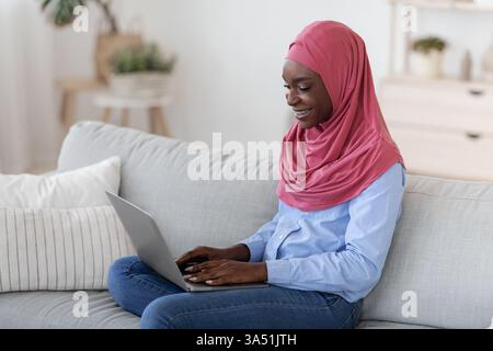 Die Millennial Black Muslim Frau mit Hijab arbeitet zu Hause ferngesteuert auf einem Laptop und genießt freiberufliche Arbeit von einer gemütlichen Couch aus. Dieses Lifestyle-Bild zeigt Remote-Arbeit, Home Office-Einrichtung und flexible Karrieren für verschiedene Profis. Ideal für Artikel über Freiberufler, digitale Nomaden und integrative Arbeitsplätze. Stockfoto