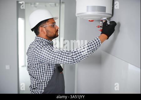 Indischer männlicher Klempner in Uniform und Schutzhelm, der an einem Heizsystem im Badezimmer arbeitet. Ein professioneller Reparaturspezialist mit Fokus auf HLK. Geeignet für Hausverbesserungen, Sanitär- und Heizungswartungskampagnen. Stockfoto