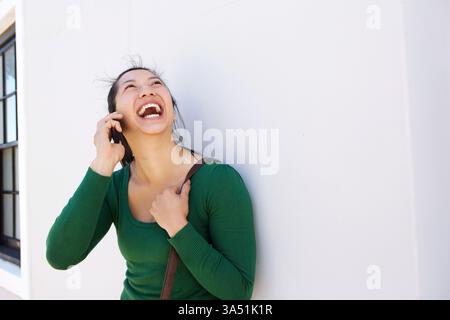 Fröhliche asiatische Frau bei einem Handy, die draußen mit einem Taschenriemen an einer weißen Wand steht. Dieses moderne Lifestyle-Bild erfasst eine sichere Kommunikation für geschäftliche und private Zwecke. Geeignet für Lifestyle, Mobiltechnologie und Kommunikationskampagnen. Stockfoto