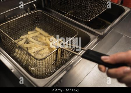 Eine Hand verwendet ein Metallwerkzeug, um goldene Pommes Frites aus einer Fritteuse zu heben. Die Küche ist geschäftig, voll mit den Klängen und Aromen von Speisen, die zubereitet werden Stockfoto