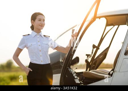 Chinesischer Hubschrauberpilot steht neben einem Hubschrauber auf einem Flugplatz. Die Vorderansicht und die hintergrundbeleuchtete Beleuchtung erzeugen ein dramatisches, professionelles Porträt, das Fachwissen und Ehrgeiz in der Luftfahrt unterstreicht. Ideal für die Luft- und Raumfahrtindustrie und Frauen in MINT-Kampagnen. Stockfoto