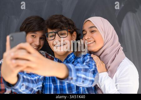 Gruppe arabischer Teenager, die Selfies mit einem Smartphone vor einem schwarzen Tafel machen. Der selektive Fokus hebt einen lebendigen, geselligen Moment unter muslimischen Jugendlichen hervor. Perfekt für Inhalte zu Jugend, sozialen Medien und Vielfalt in Gemeinden im Nahen Osten. Stockfoto