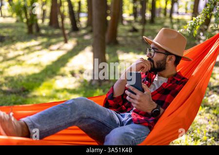 Bärtiger Mann mit Strohhut entspannt sich draußen in einer Hängematte. Die ruhige Sommerszene vermittelt Ruhe und Freizeit, ideal für Reise- und Wellnesskampagnen. Stockfoto