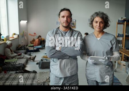 Zwei selbstbewusste Ärzte oder Freiwillige in medizinischen Peelings, die in die Kamera schauen, während sie in einem geräumigen Raum für Flüchtlinge stehen Stockfoto