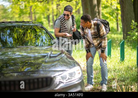 Verschiedene Gruppen von Freunden, Schwarze und Kaukasier, unterhalten sich an einem sonnigen Tag mit dem Fahrer auf einer Forststraße. Diese Outdoor-Szene vermittelt Abenteuer, Freiheit und lässige Kameradschaft am Straßenrand, während sie per Anhalter in die Landschaft und den Park wandern. Es ist ein lebendiges Bild für Themen wie Freundschaft, Roadtrips und Sommerurlaub. Stockfoto