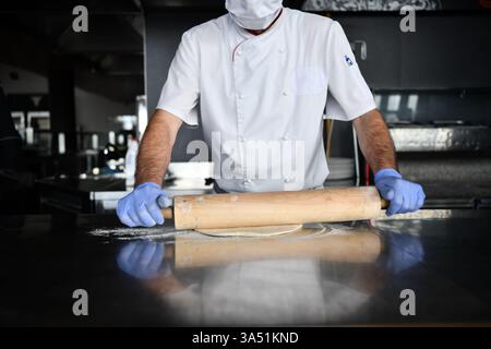 Der erfahrene Koch in einer modernen Restaurantküche bereitet traditionelle italienische Pizza im Holzofen zu. Das Tragen einer Maske als Teil der Sicherheitsmaßnahmen verbindet professionelles Kochen mit authentischem Backofen-Ambiente. Ideal für Gastronomie, Pizza und Dining Visuals. Stockfoto