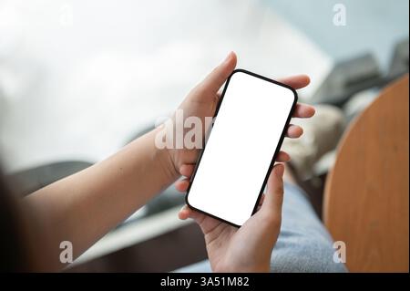 Draufsicht einer Frau, die ein Smartphone mit einem leeren weißen Bildschirm hält und an einem Tisch in einem Café sitzt. Dieses vielseitige technische Image eignet sich für Apps, Mobilgeräte, Remote-Arbeiten und digitale Lifestyle-Themen. Stockfoto
