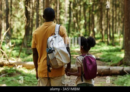 Rückansicht einer zweiköpfigen Familie mit Rucksäcken, die an einem sonnigen Sommertag durch einen Wald wandern. Die Szene vermittelt Outdoor-Abenteuer und Naturerkundungen, die für Reisen und Familienaktivitäten geeignet sind. Ideal für Geschichten über Wandern, Binden und Wochenendausflüge im Freien. Stockfoto