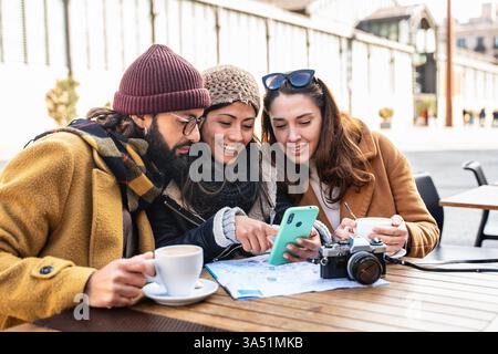 Junge Freunde, die während des Winterurlaubs in einem Café auf ein Smartphone schauen, während sie sich einen Stadtplan ansehen. Vielseitiges urbanes Lifestyle-Image, ideal für Reiseplanung, Städtereisen und Gruppenabenteuer. Ideal für Kampagnen, die sich auf Freundschaft, Erkundung und Winterreisen konzentrieren. Stockfoto
