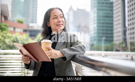 Eine selbstbewusste, reife asiatische Geschäftsfrau steht auf einem Skywalk im Freien und hält ein Buch und einen Kaffee zum Mitnehmen in der Hand. Sie strahlt Führungsstärke und Ehrgeiz in einer Stadt aus. Perfekt für Führungs-, Mentoring- und Erfolgskampagnen. Stockfoto