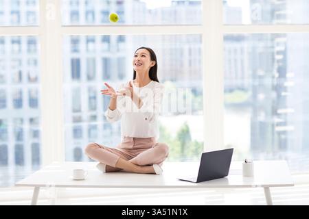 Die lächelnde chinesische Geschäftsfrau in ihren 20ern wirft einen grünen Apfel in die Luft neben einem Laptop auf einem modernen Schreibtisch. Der lebendige Moment vermittelt Selbstvertrauen und eine positive, produktive Arbeitsstimmung. Perfekt für den Unternehmensstil, die Bürokultur und gesunde Arbeitszeiten. Stockfoto