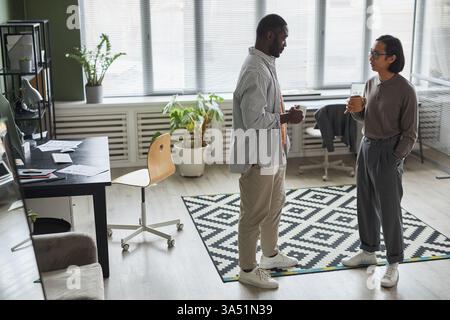 Zwei verschiedene Fachleute, ein Afroamerikaner und ein asiatischer Kollege, teilen sich Kaffee in einem modernen Büro. Sie unterhalten sich und arbeiten zusammen und veranschaulichen Teamarbeit, Networking und gelegentliche Geschäftsinteraktionen mit Kopierflächen. Stockfoto