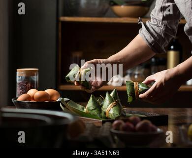 Nahaufnahme der Hände, die Zongzi für das Dragon Boat Festival herstellen. Ein traditionelles chinesisches Gericht, das in der Küche mit Blättern und frischen Zutaten zubereitet wird. Stockfoto