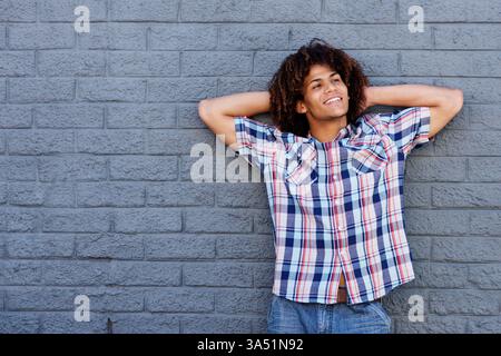 Lächelnder Afroamerikaner mit Afro-Frisur, Hände hinter dem Kopf, lehnt sich an eine Ziegelmauer auf der Straße. Lässige, unbeschwerte Atmosphäre macht dieses Bild ideal für Lifestyle, urbane Mode und moderne Porträts. Ideal für positive, positive Kampagnen und Inhalte. Stockfoto