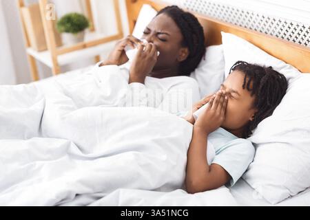 Schwarze Mutter und Tochter im Bett, unwohl und niesen, während sie mit Papierservietten Nasen blasen. Eine realistische Gesundheitsszene für Familien-, Gesundheits- und Erkrankungskampagnen. Geeignet für Artikel über Atemwegsinfektionen, Grippesaison und häusliche Pflege. Stockfoto