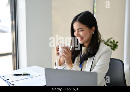 Die charmante asiatische Geschäftsfrau der Millennials genießt einen Morgenkaffee an ihrem Schreibtisch in einem modernen Büro. Dieses fröhliche Porträt hebt die Themen des beruflichen Lebensstils, des Marketings und der Führung hervor. Ideal für Unternehmenskommunikation und Kampagnen am Arbeitsplatz. Stockfoto