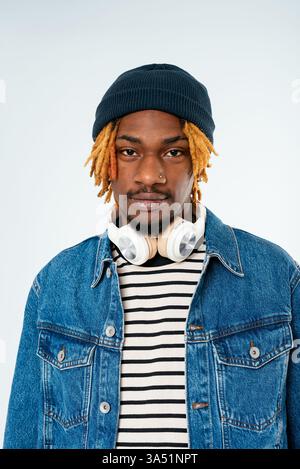 Ernster Schwarzer Mann mit Dreadlocks, der eine Mütze und Kopfhörer um den Hals trägt, vor weißem Hintergrund. Ideal für Musik, Mode oder urbane Stilkonzepte. Starkes, selbstbewusstes Porträt mit einem modernen, legeren Look. Stockfoto