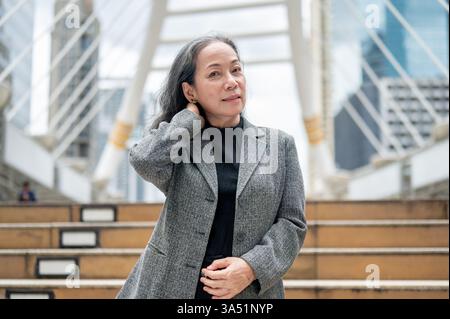 Asiatische Geschäftsfrau in einem scharfen Anzug steht auf einem Skywalk über einem modernen Stadtbild. Sie blickt selbstbewusst in die Kamera und verkörpert Führungsstärke und Erfolg. Ideal für geschäftliche, Banking- oder unternehmerische Funktionen. Stockfoto
